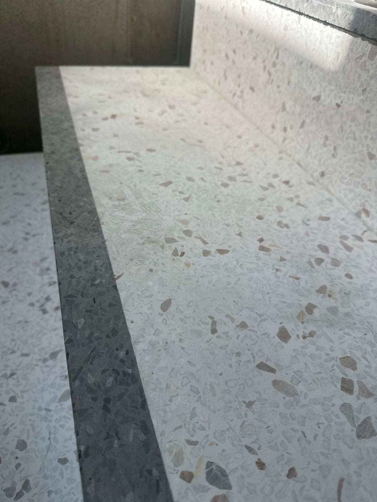 Nærbilde av lyst terrazzogulv med grå og beige steinsplitt, en mørkere grå kantlist og oppbrett i terrazzo mot vegg