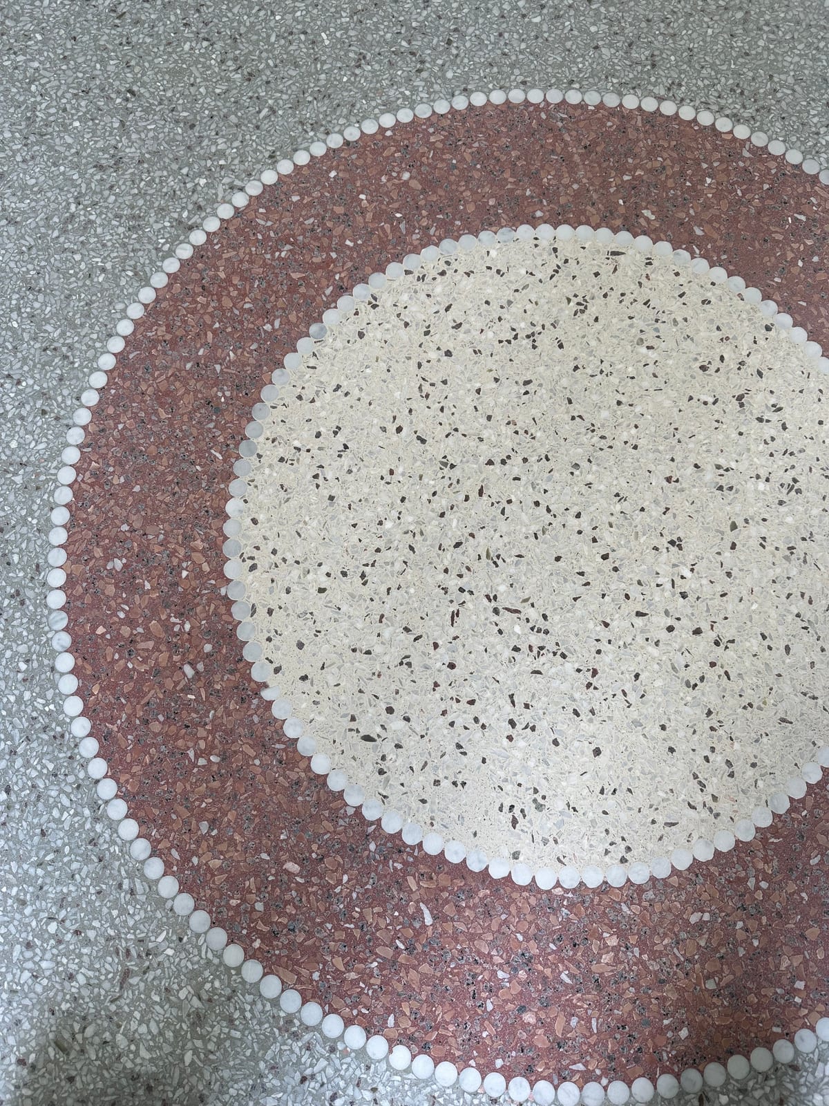 Sirkelmotiv i terrazzo med lys kjerne, rødbrun ring og rammer av hvite steinperler i grått gulv